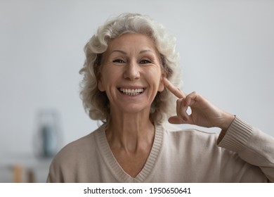 Portrait of happy senior woman pointing finger at her ear, looking at camera and smiling. Excited mature 60s lady enjoying hearing ability after deafness therapy. Deaf patient communication. Head shot