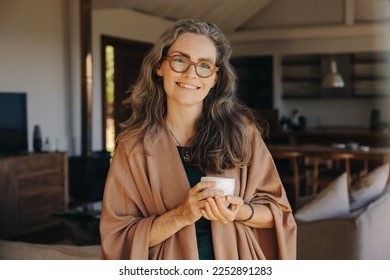 Portrait of a happy senior woman smiling at the camera while standing with a cup of tea in her hands. Cheerful senior woman enjoying a serene retirement at home. - Powered by Shutterstock