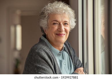 Portrait of happy senior woman standing at home near window and looking at camera. Smiling beautiful old woman relaxing at home during Covid quarantine. Cheerful retired old lady relaxing at home.