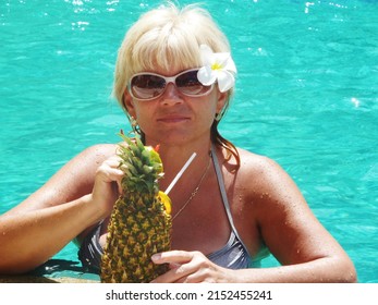 Portrait Of Happy Senior Woman In Swimming Pool Drinking Exotic Cocktail In Pineapple Fruit. Beautiful Glamour Mature Woman Drinking Cocktail In Luxury Pool Looking At Camera. Enjoying Summer Vacation