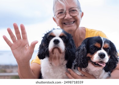 Portrait of happy senior woman in yellow jersey waving hand sitting close to the beach with her two cavalier king charles dogs. Best friend forever concept - Powered by Shutterstock