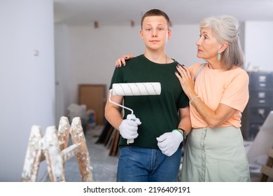 Portrait Of Happy Senior Woman And Young Man With Paint Roller. Teenager Working Construction Site During Vacation. Grandmother Proud Of Her Grandson Assisting With Home Improvement Works.