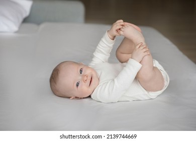 Portrait of happy sincere cute few months baby boy girl holding tiny feet in hands, lying alone on cozy bed, having fun playing after waking up, looking at camera, carefree childhood concept.