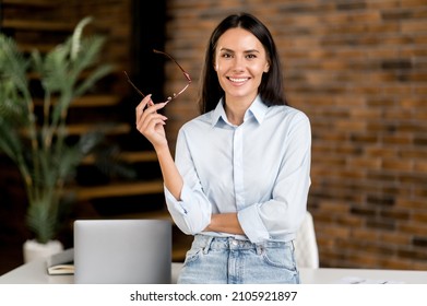 Portrait happy smart successful caucasian brunette business woman, ceo, top manager, real estate agent, stands near table in modern office, in stylish clothes, looks at camera, smiles friendly - Shutterstock ID 2105921897
