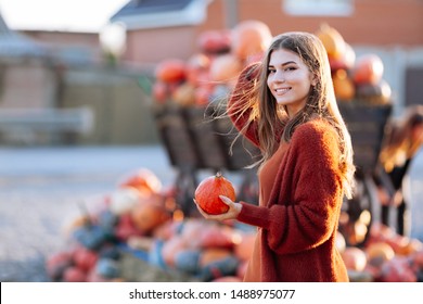 Portrait of happy smile woman with small pumpkin in hand near wooden wagon with pumpkin on farmers market in brown sweater, dress and hat. Cozy autumn vibes Halloween, Thanksgiving day.