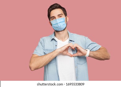 Portrait of happy smiley young man with surgical medical mask in blue shirt standing with love hand gesture on heart and looking at camera smiling. indoor studio shot, isolated on pink background.
