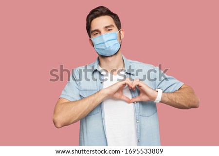 Portrait of happy smiley young man with surgical medical mask in blue shirt standing with love hand gesture on heart and looking at camera smiling. indoor studio shot, isolated on pink background.