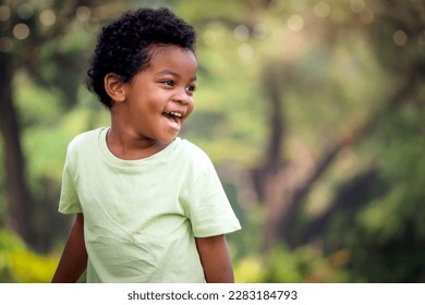 Portrait of happy smiling African boy with black curly hair standing and laughing outdoor with blurred green garden background, beautiful kid play outside park on sunny day, cute child play in summer. - Powered by Shutterstock