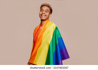 Portrait of happy smiling afro latin american young man with a gay pride rainbow flag at studio over beige background. Homosexual lgbtiq concept, rainbow flag, celebrating parade. Copy space.