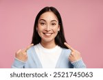 Portrait of happy smiling Asian woman pointing at her perfect white teeth smiling isolated on pink background, looking at camera. Dental care, treatment, health care concept