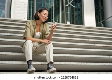 Portrait of happy smiling asian woman, sitting outdoors near building, using smartphone. Technology concept. - Powered by Shutterstock