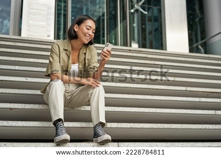 Portrait of happy smiling asian woman, sitting outdoors near building, using smartphone. Technology concept.