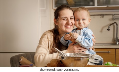 Portrait Of Happy Smiling Baby Boy Covered In Flour And Dough With Smiling Mother Posing On Messy Kitchen After Baking Bread Or Cake