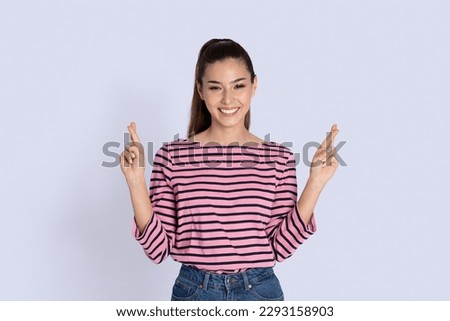 Portrait of happy smiling beautiful young woman with long brunette hair standing over grey studio background, holding fingers crossed for good luck and smiling at camera, copy space