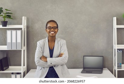 Portrait of happy smiling black businesswoman at work. Beautiful young African American business woman in jacket and glasses standing arms folded leaning on table in office with grey copy space wall
