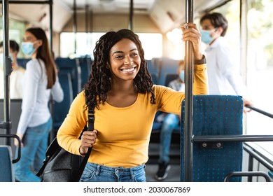 Portrait of happy smiling black female without mask standing inside public transportation with backpack, looking away out of window, group of people in facemask walking out in blurred background