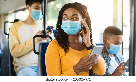 Portrait of happy smiling black female in medical face mask traveling on public transit, listening to music touching wireless earphones and using cellphone, looking away at window, sitting inside - Shutterstock ID 2030067971