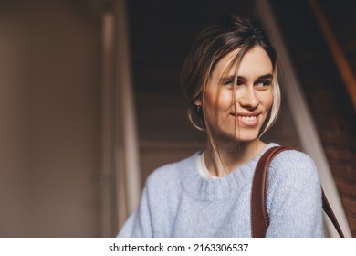 Portrait Of Happy Smiling Blonde Woman Exit Her Home She Go Away With Bag On Shoulder. Blonde Happy Woman With Low Bun Wear Blue Sweater. Young And Beautiful Girl Look At Side.