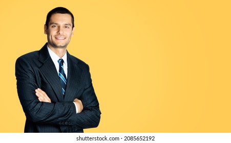 Portrait Of Happy Smiling Brunette Businessman In Black Suit, White Shirt And Blue Stripes Tie Standing In Crossed Arms Pose Over Orange Yellow Color Background. Confident Business Man. Male Executive