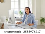 Portrait of a happy smiling business woman accountant sitting at the desk and working on a pc computer with tables and charts at modern office or at home and looking at the camera.