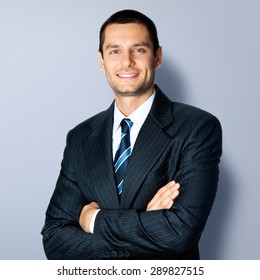 Portrait Of Happy Smiling Businessman In Crossed Arms Pose, In Black Confident Suit, Against Grey Background. Caucasian Male Model At Studio Shot. Business And Success Concept.