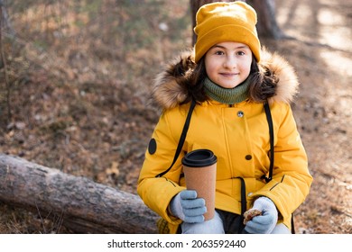 Portrait of a happy smiling caucasian girl in orange clothes in the autumn pine forest. Attractive teenager sits resting and drinking a hot beverage. Outdoor activity. Sunny autumn day. Copy space