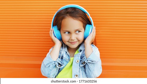 Portrait of happy smiling child in headphones listening to music on city street over orange wall background
