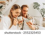 Portrait of happy, smiling couple drinking coffee holding cup with drink, sitting on comfortable sofa in living room at home. Morning routine, relationship concept