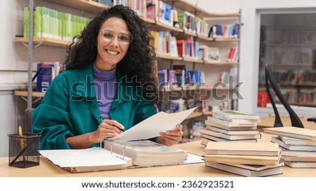 Portrait of a happy smiling curly girl with glasses sitting in library,  Bookshelves background, sorting out papers working checking textbooks accepting or issuing. 
Buying a book at a bookstore. 
