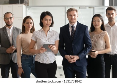 Portrait Of Happy Smiling Diverse Employees Team Standing In Office, Holding Phone And Tablet, Posing For Company Photo Together, Looking At Camera, Workers Group Motivated For Business Success