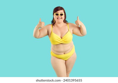 Portrait of happy smiling fat woman wearing yellow swimsuit showing thumb up sign isolated on a blue studio background. Travelling, body positive, vacation and summer journey concept.