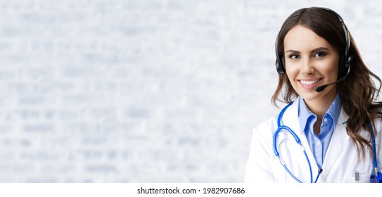 Portrait Of Happy Smiling Female Doctor In Headset, Against White Brick Loft Wall Background, With Empty Copy Space Area. Medical Call Center Concept Picture. Zoom Online Consulting.