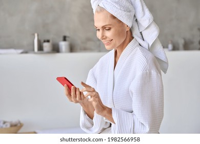 Portrait Of Happy Smiling Gorgeous Middle Aged Lady Wearing Bathrobe And Turban Towel On Head Using Mobile Beauty Apps Holding Smartphone. Antiage Skin And Hair Care Spa Procedures Concept.