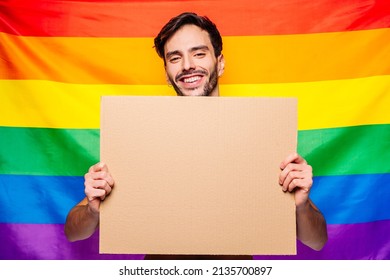 Portrait Of Happy Smiling Homosexual Man Holding Empty Blank Board, With A Gay Pride Flag Behind Him At Studio. Lgbtq Flag, Rainbow Flag, Celebrating Parade.