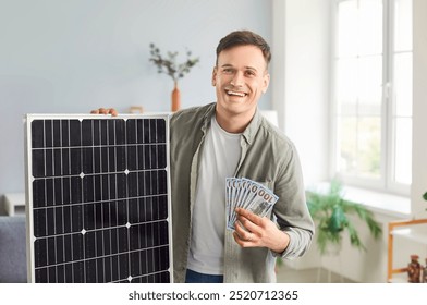 Portrait of a happy, smiling, joyful man at home holding a photovoltaic solar panel and a fan of US dollar bills. Concept of using sustainable technology and clean solar energy to save money - Powered by Shutterstock