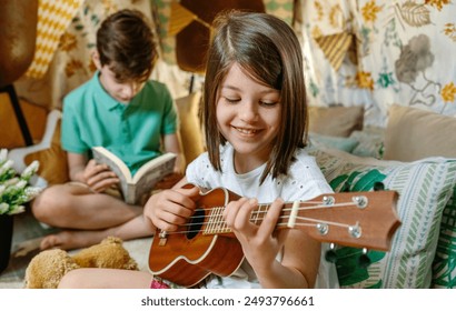 Portrait of happy smiling little girl playing ukulele while boy reading book on handmade shelter tent. Children having fun in diy teepee in their house. Vacation camping at home or staycation concept. - Powered by Shutterstock