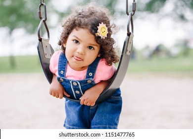 Portrait Of Happy Smiling Little Latin Hispanic Toddler Girl Swinging On Swings At Playground Outside On Summer Day. Happy Childhood Lifestyle Concept. Toned With Film Pastel Faded Filters Colors.