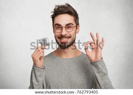 Portrait of happy smiling male with bristle shows ok sign as holds condomn, agrees to spend night with beautiful woman. Caucasian bearded guy advertises new kind of contraceptive. Safety and health