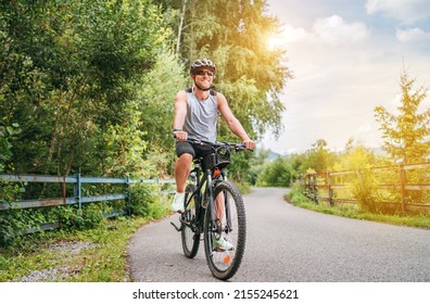Portrait of a happy smiling man dressed in cycling clothes, helmet and sunglasses riding a bicycle on the asphalt out-of-town bicycle path. Active sporty people concept image. - Shutterstock ID 2155245621