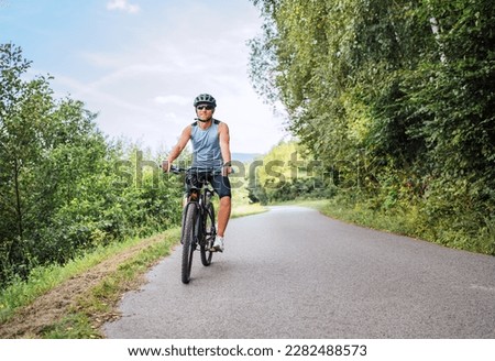 Portrait of a happy smiling man dressed in cycling clothes, helmet and sunglasses riding a bicycle on the asphalt out-of-town bicycle path. Active sporty people concept image. 