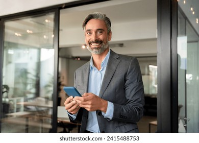 Portrait of happy smiling mature European latin businessman holding smartphone in office, copy space. Middle aged man using cell phone mobile app and looking at camera. Applications for business. - Powered by Shutterstock