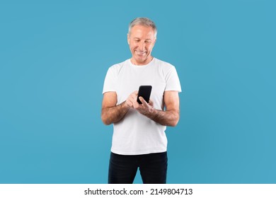 Portrait Of Happy Smiling Mature Man Using Smartphone Standing Isolated Over Blue Studio Background, Copy Space. Adult Male Chatting Online, Browsing Social Media. Older People And Technology Concept