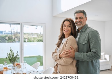 Portrait of happy smiling mature older family couple real estate owners standing at home, affectionate senior middle aged man and woman in love hugging in modern house house living room interior.