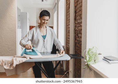 Portrait of happy smiling mature woman ironing her clean dry blouse, preparing her outfit to go to party on evening, pressing electric wired hot iron gently to board. Household chores concept - Powered by Shutterstock