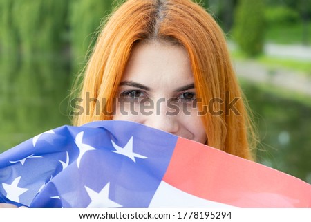 Portrait of happy smiling red haired girl hiding her face behind USA national flag. International day of democracy concept.