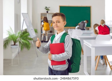 Portrait of a happy smiling schoolboy showing thumb up . Portrait of cheerful boy with backpack on his shoulders and and textbook in hand looking at the camera with classmates in background. - Powered by Shutterstock