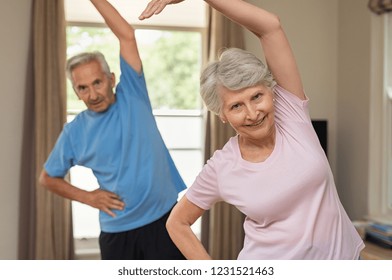 Portrait Of Happy Smiling Senior Couple With Arms Raised Doing Stretching Exercise At Home. Old Man And Elderly Woman Practicing At Home During Early Morning. Retired Couple Doing Aerobics Workout.