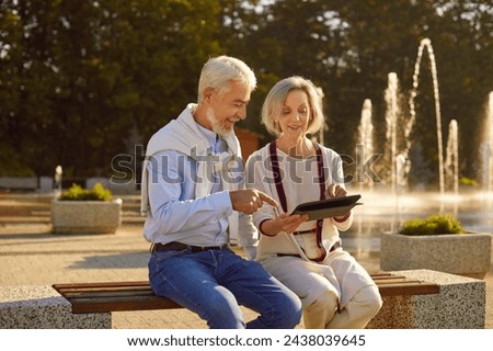 Portrait of happy smiling senior couple sitting outdoors on bench in city park using digital tablet for social media, searching information, watching funny movie or having online video call.