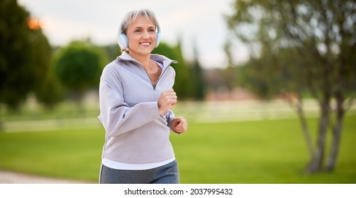 Portrait of happy smiling senior lady in sportswear jogging in early morning, wearing wireless cordless headphones, running in city park along green trees. Healthy and active lifestyle concept