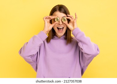 Portrait of happy smiling woman covering eyes with gold bitcoin, blockchain concept, cryptocurrency, digital money, wearing purple hoodie. Indoor studio shot isolated on yellow background.